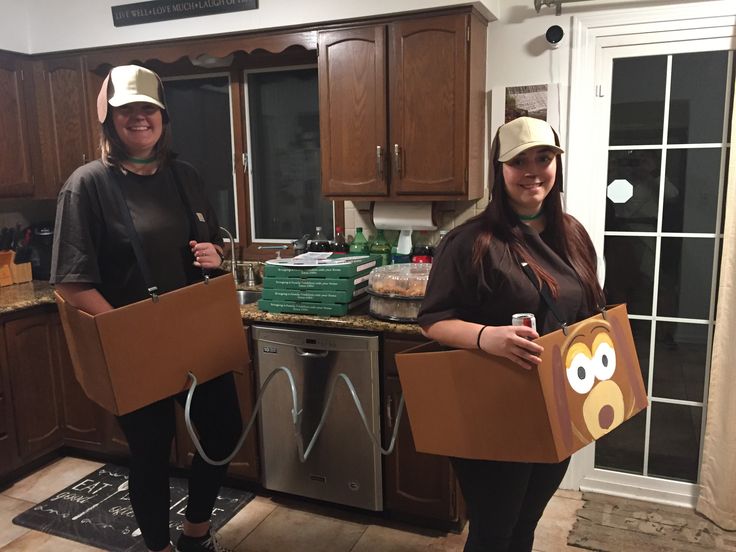 two women standing in a kitchen holding cardboard boxes with faces on them and one has a bear face on it