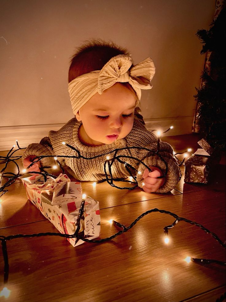 a baby is sitting on the floor with christmas lights around her and looking at a present