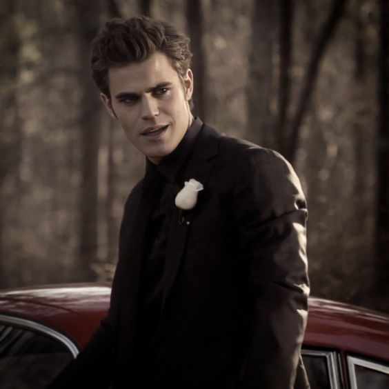 a young man standing next to a red car in the woods with trees behind him