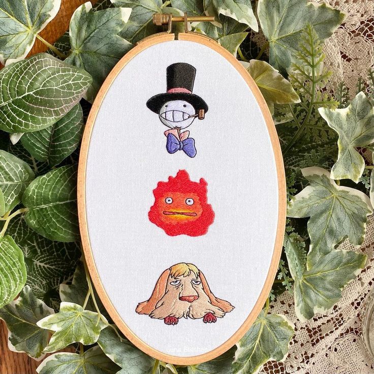 the embroidery is hanging on the wall next to some green plants and leaves, which have been decorated with cartoon characters