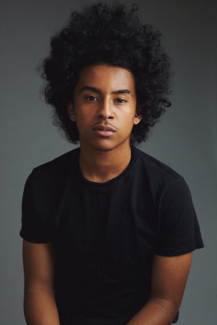 a young man with an afro sitting in front of a gray background wearing a black t - shirt