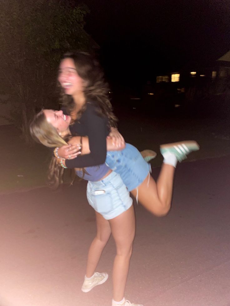 two young women are hugging each other on the street at night, one is wearing white shoes and the other has blue denim shorts