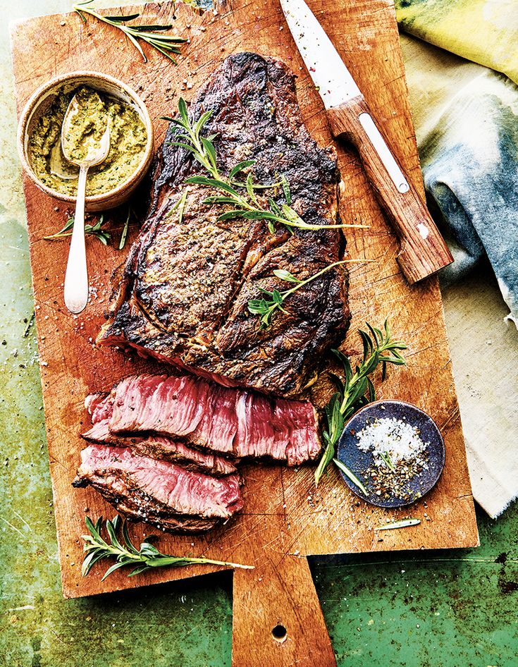 a steak on a cutting board with herbs and seasoning next to it, along with two small bowls of sauces