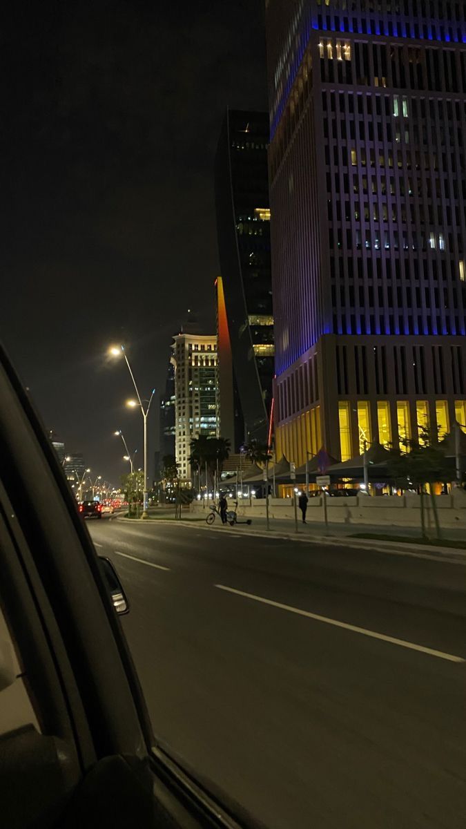 a city street at night with tall buildings in the background and cars driving on the road