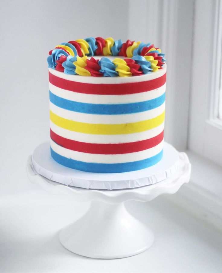 a multicolored cake sitting on top of a white cake stand next to a window