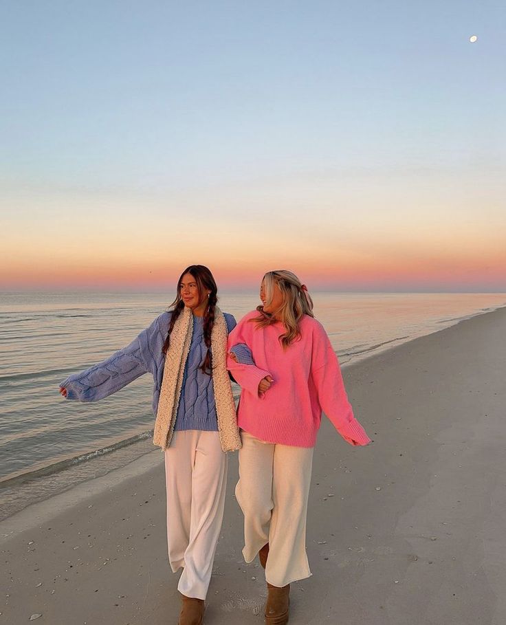 two women walking on the beach at sunset with their hands in each others'pockets