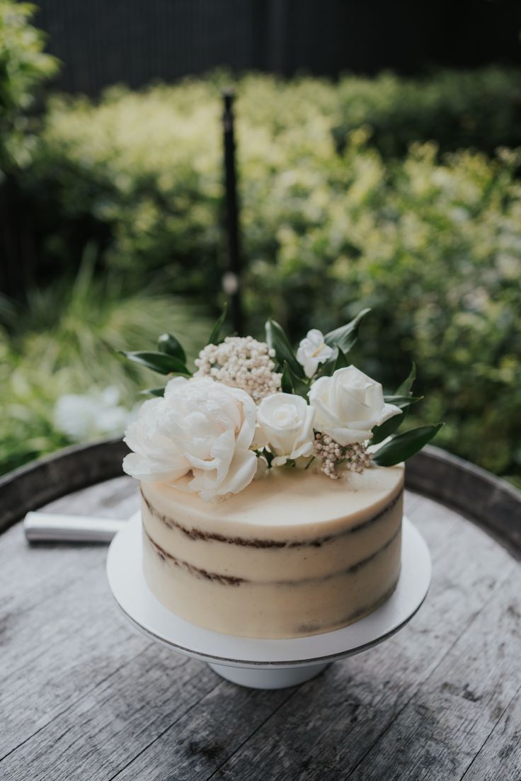 a white cake with flowers on top sitting on a wooden table in front of some bushes