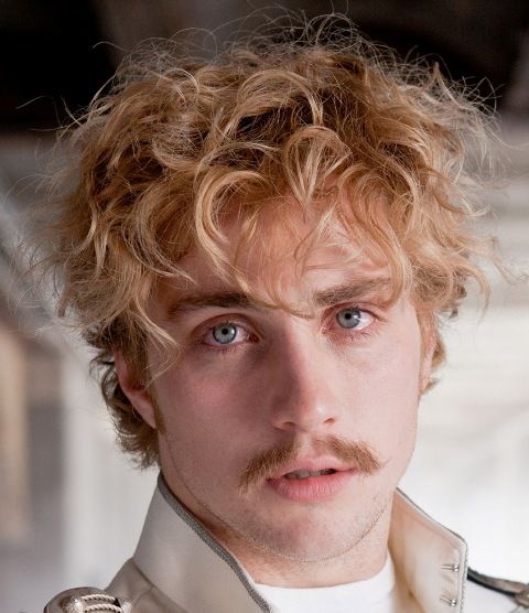 a man with curly hair and a moustache looks at the camera while wearing a jacket