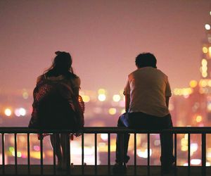 two people sitting on top of a metal fence overlooking a city at night with lights in the background