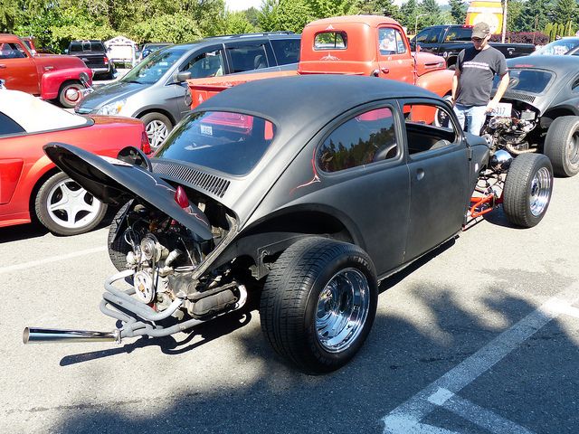 an old car is parked in a parking lot with other cars behind it and people standing around