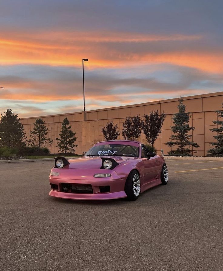 a pink car parked in a parking lot next to a tall building with the sun setting behind it