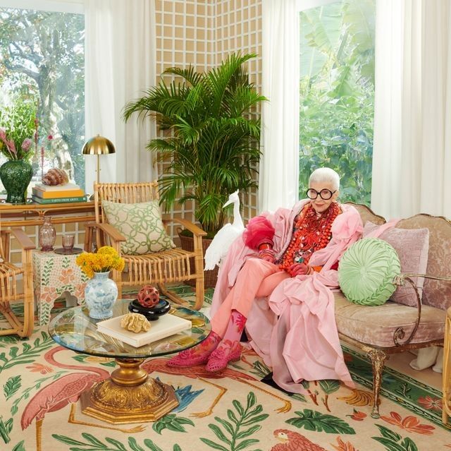 an old woman sitting on a couch in a living room with plants and flowers around her