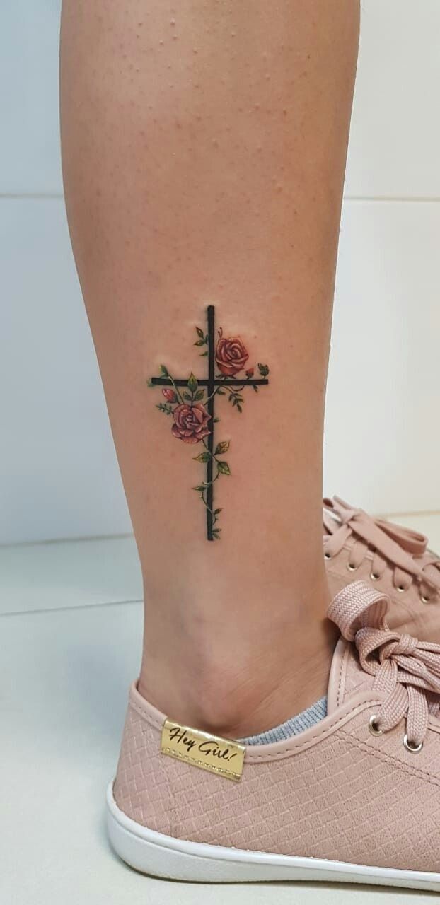 a small cross tattoo on the ankle with pink roses and green leaves around it's edges