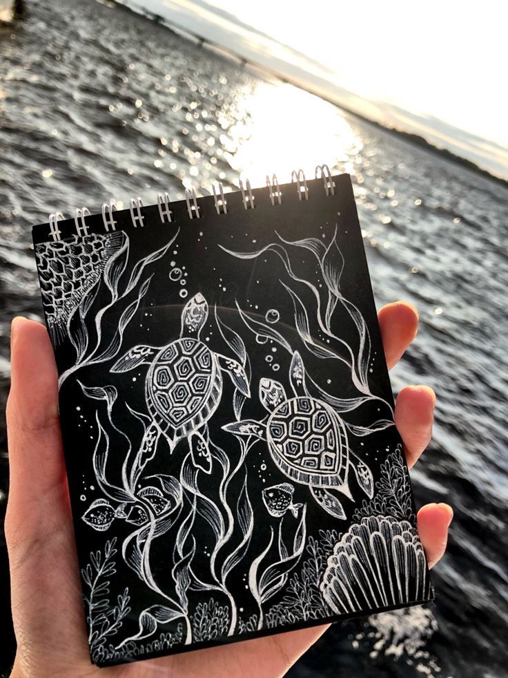 a hand holding up a black and white notebook with sea animals on it in front of the ocean