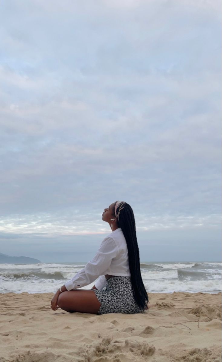 a woman sitting on top of a sandy beach next to the ocean and looking up