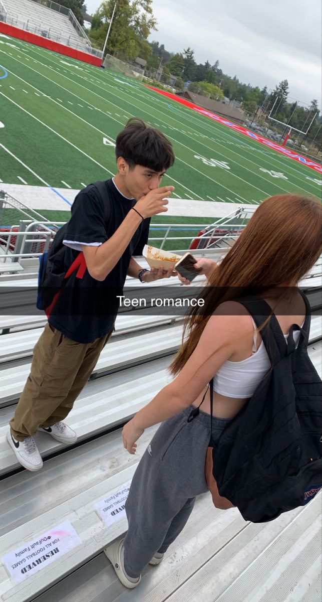 two young people standing on the bleachers at a football game, one is eating