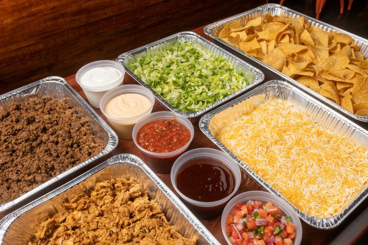 several trays of food are arranged on a table with condiments and tortilla chips