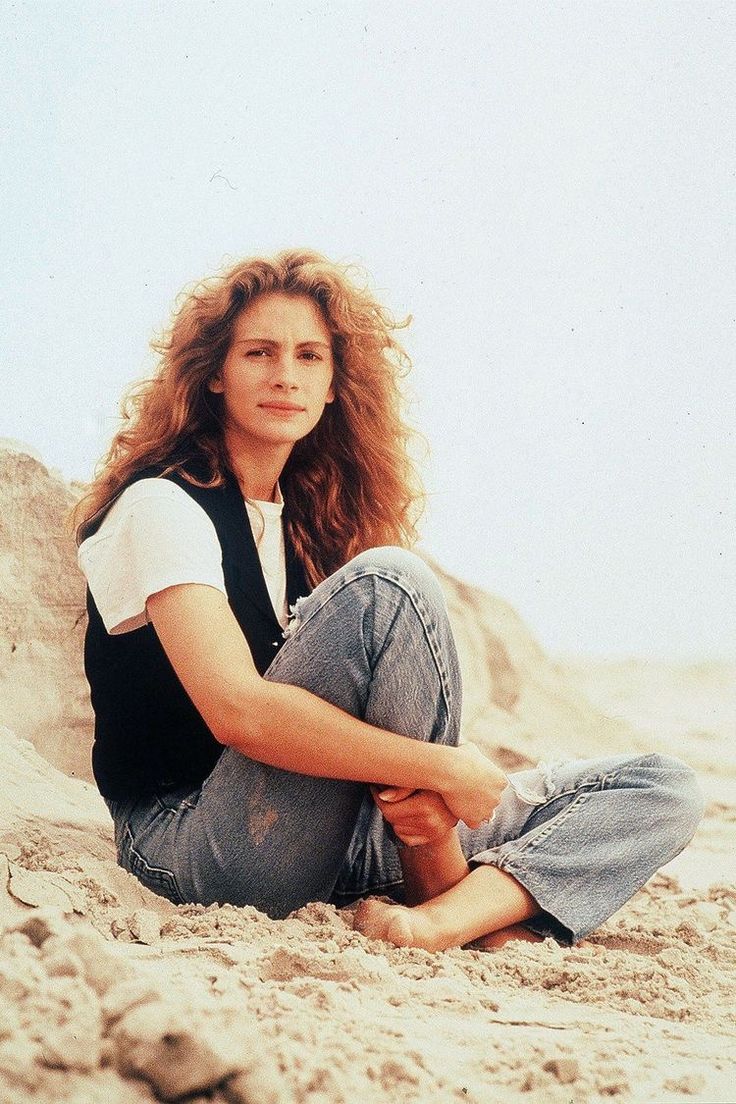 a woman sitting in the sand with her legs crossed and wearing jeans, black vest and white t - shirt