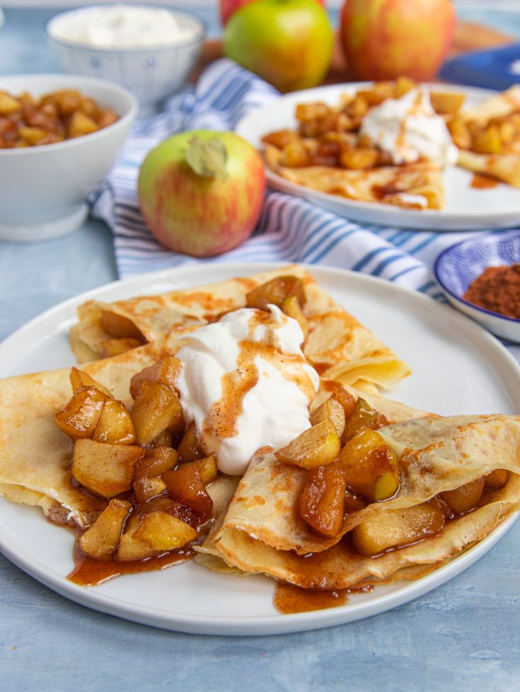 two white plates topped with crepes covered in apples and whipped cream next to bowls of fruit