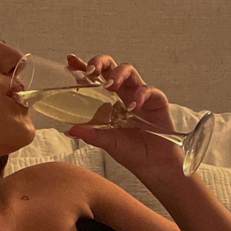 a woman drinking from a wine glass while laying on a bed with her eyes closed