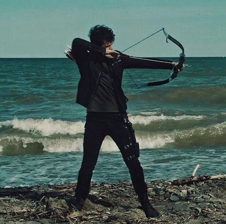 a man standing on top of a beach next to the ocean holding a bow and arrow