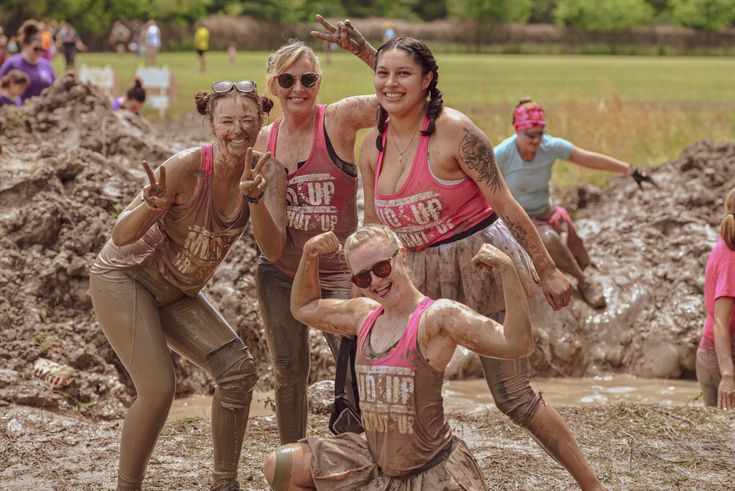 three women in mud suits are posing for a photo while other people watch from the muddy bank