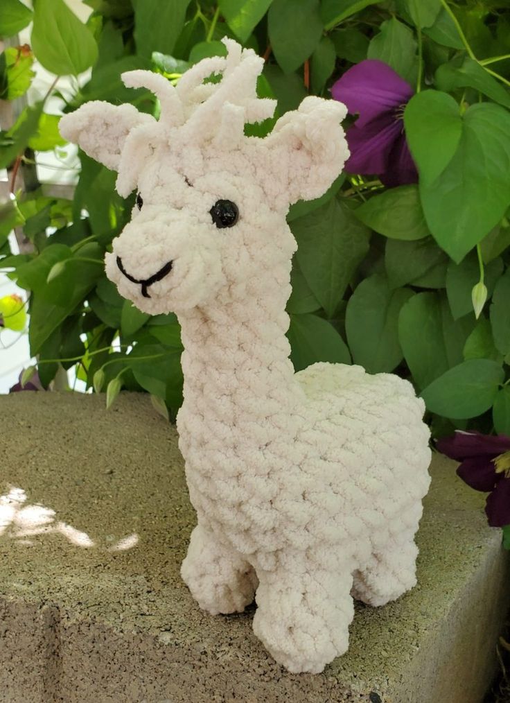 a crocheted llama sitting on top of a cement block next to flowers