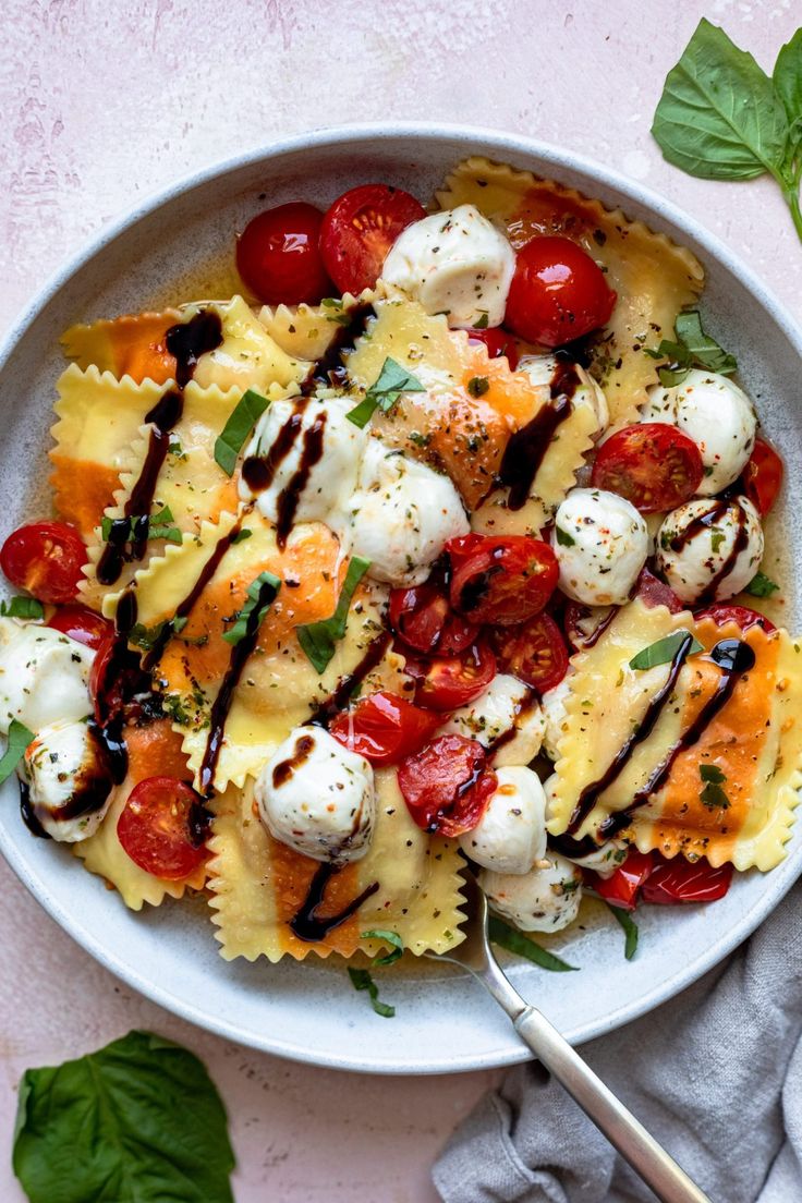 a white bowl filled with pasta covered in mozzarella and cherry tomatoes next to basil leaves