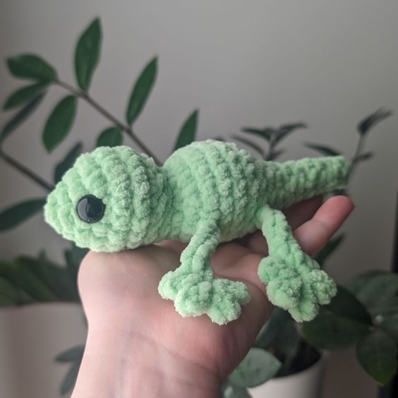 a hand holding a small green crocheted geckoe with eyes on it's face