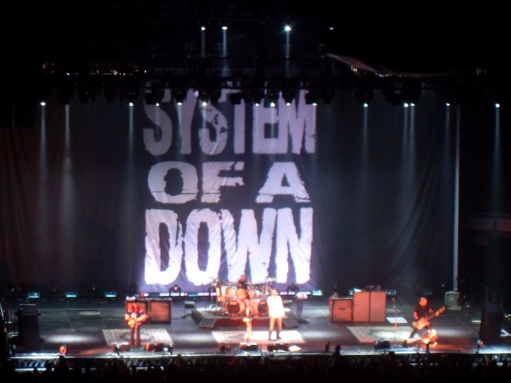 the band system performing on stage in front of a large sign that says system of a down