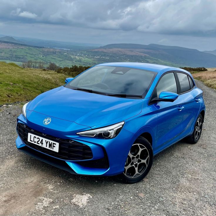 a blue car parked on the side of a dirt road next to grass and mountains