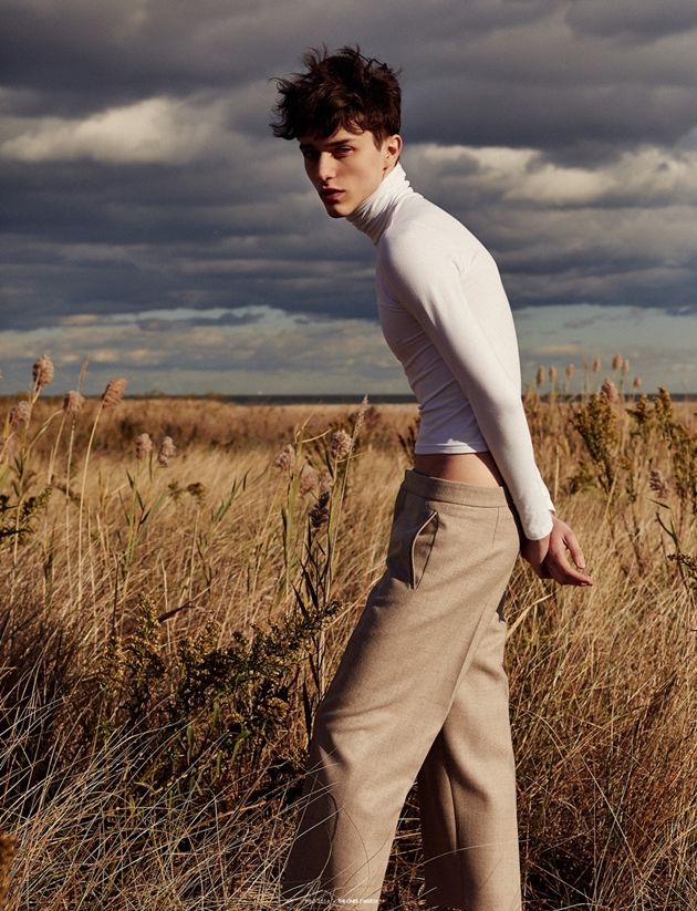 a young man standing in tall grass with his back turned to the camera, wearing white shirt and khaki pants