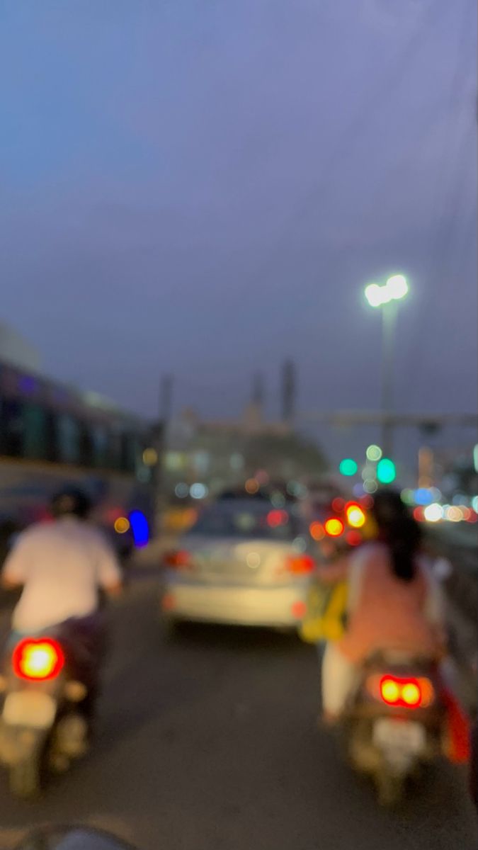 two people on motorbikes in the middle of traffic at night with cars and street lights behind them