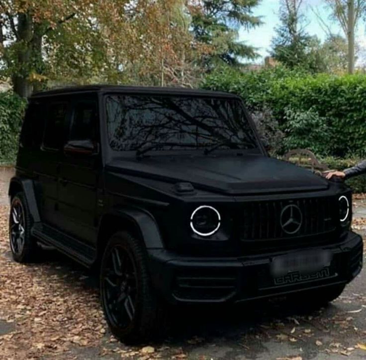 a man standing next to a black mercedes g - class in front of some trees