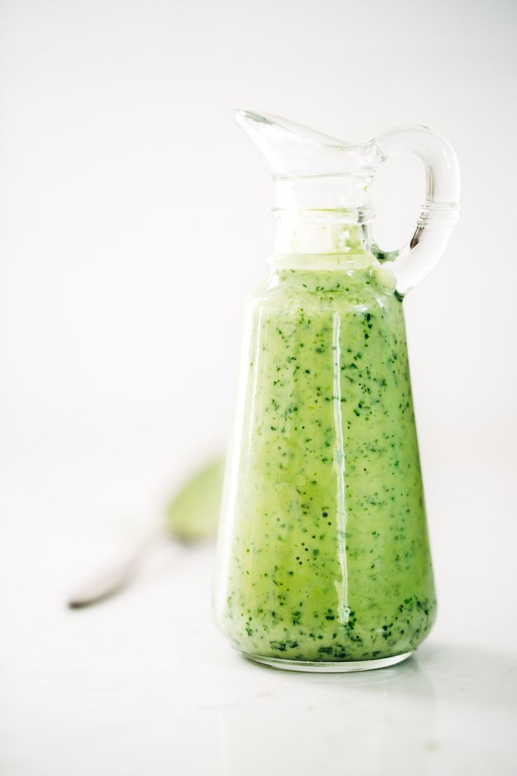 a green liquid in a glass pitcher with a spoon next to it on a white surface