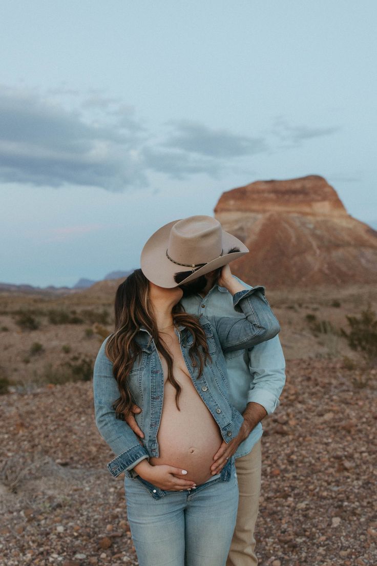 a pregnant woman standing next to a man wearing a cowboy hat in the desert at dusk