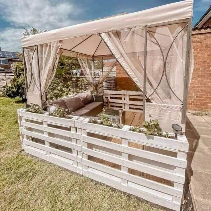 an outdoor living area with wooden pallets and white curtains on the side of it