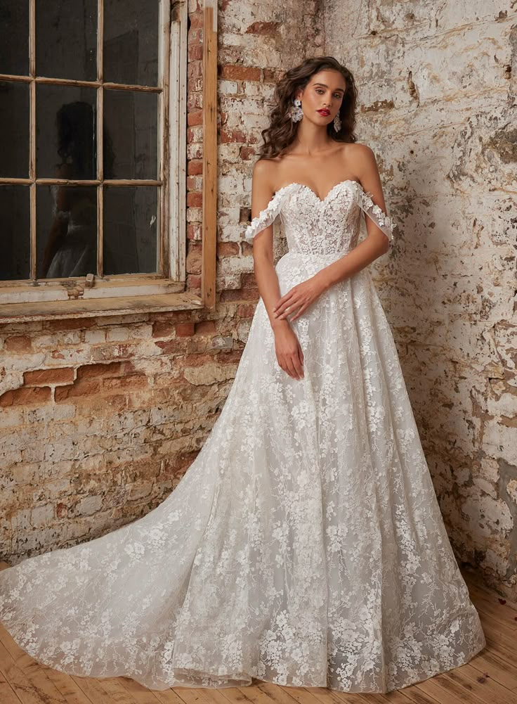 a woman standing in front of a brick wall wearing a wedding dress with off the shoulder sleeves