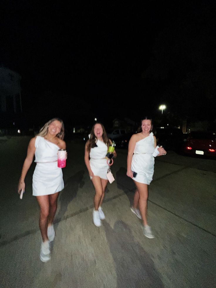 three women in white dresses are walking down the street at night, one is holding a pink cup