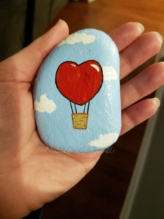 a hand holding a painted rock with a hot air balloon in the shape of a heart