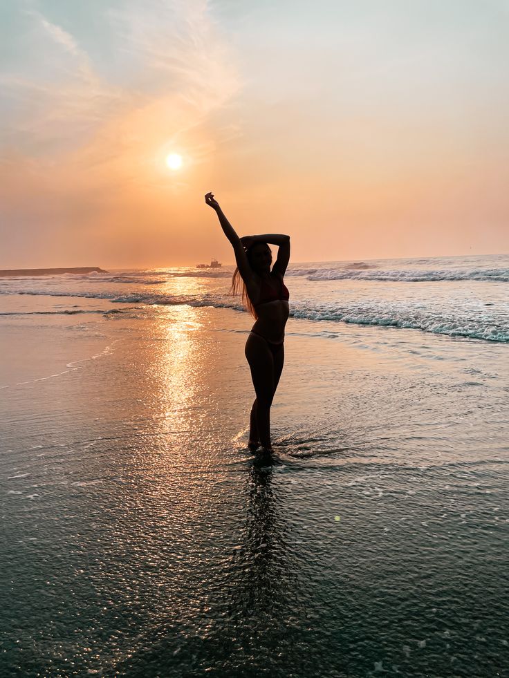 a woman is standing in the water at the beach with her arms up to the sky