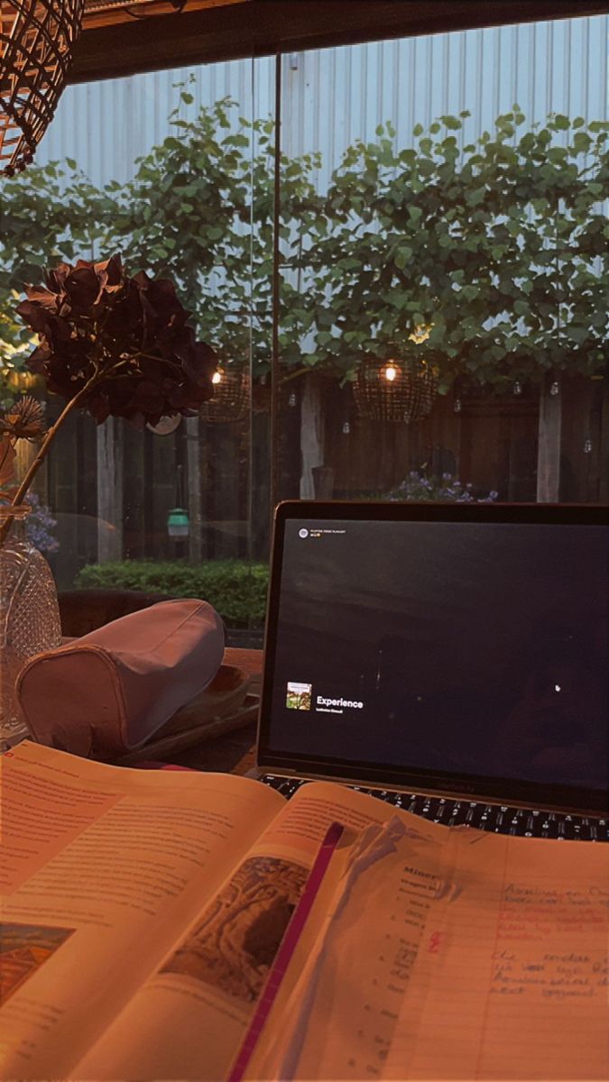 an open book sitting next to a laptop computer on top of a desk in front of a window