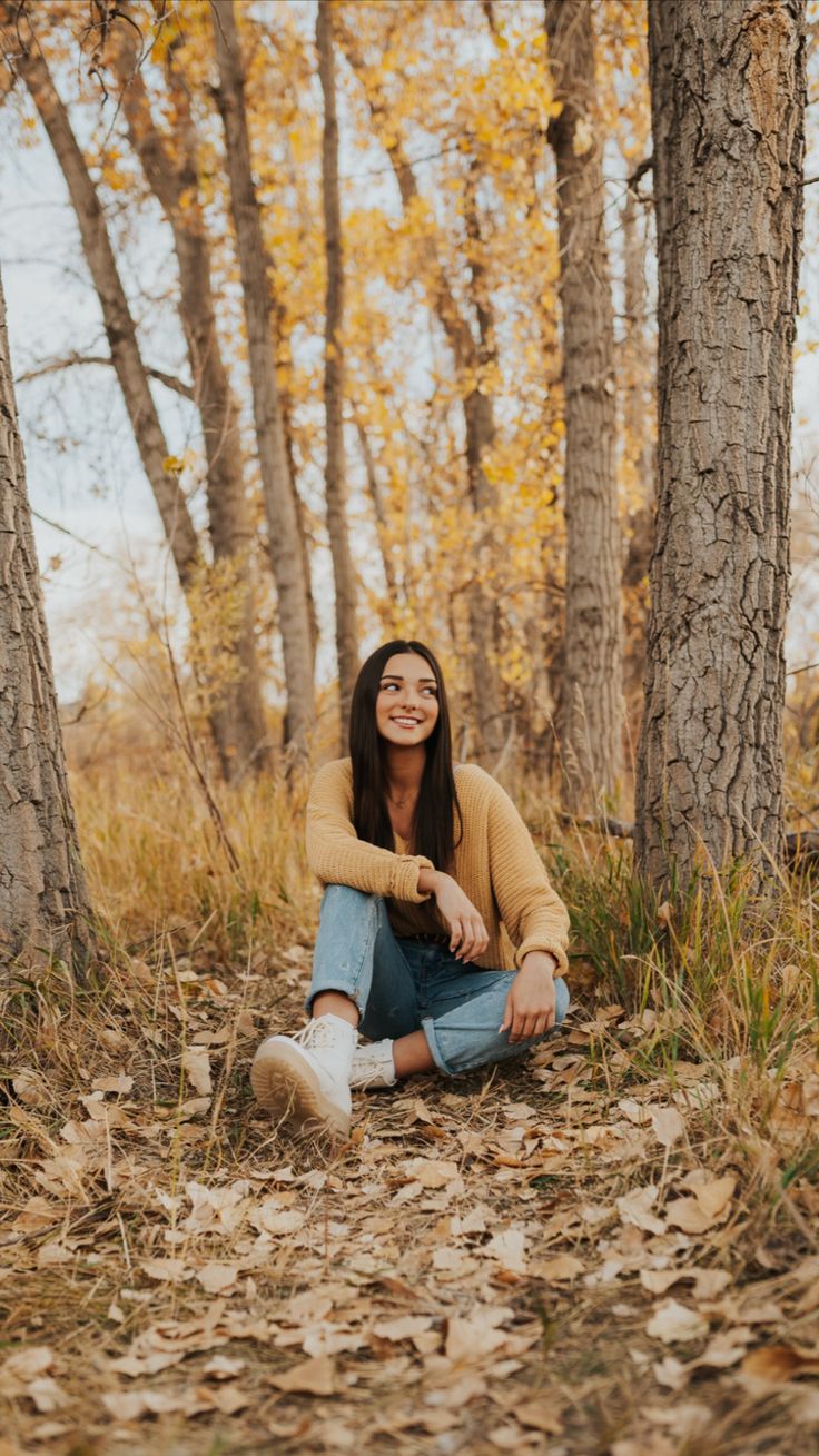 a woman sitting on the ground in front of some trees and leaves smiling at the camera