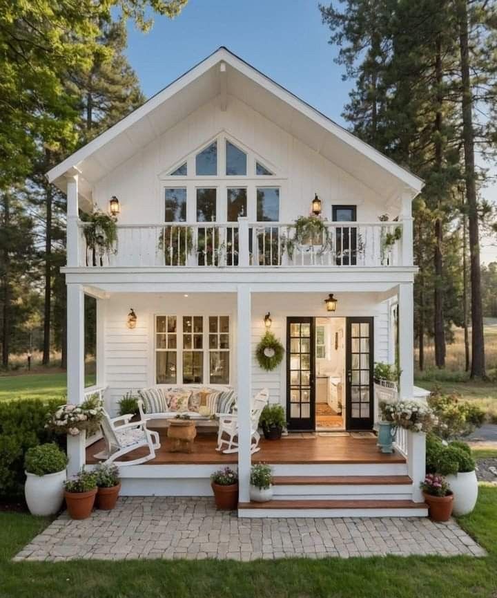 a white house with lots of windows and plants on the front porch, surrounded by greenery