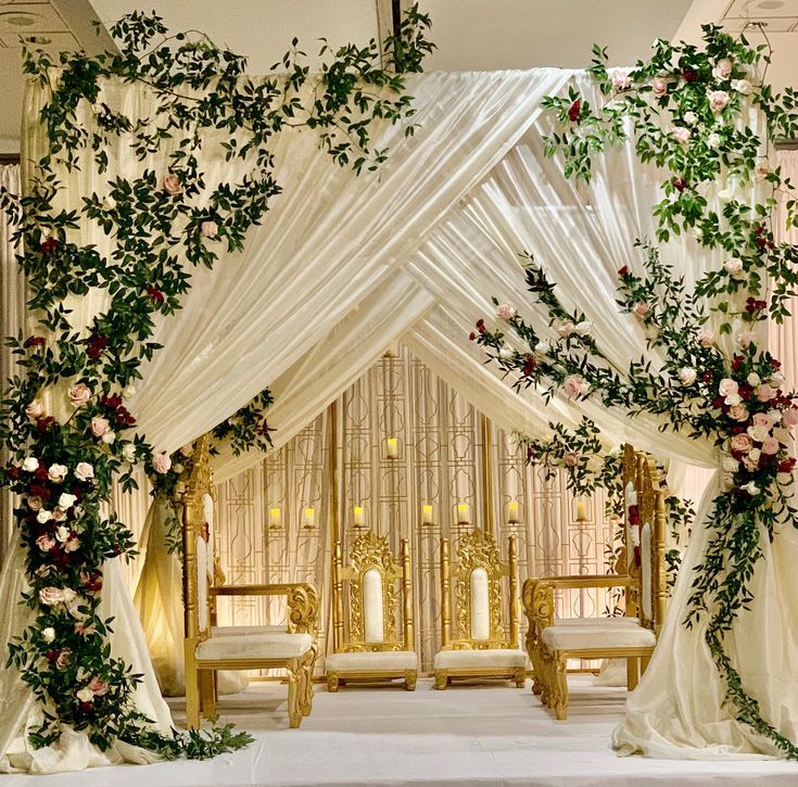 a decorated stage with white drapes and flowers on the sides, gold chairs at the end