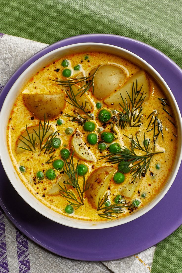 a bowl of soup with potatoes and peas on a purple plate next to a napkin