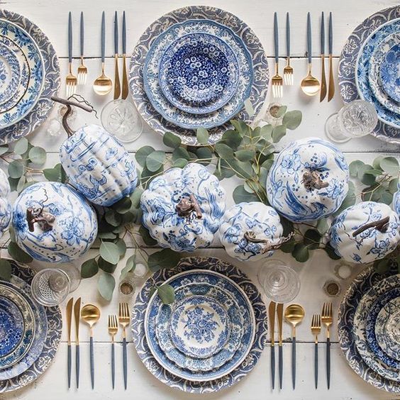 blue and white dishes are arranged on a table with gold forks, silverware and greenery