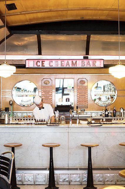 the interior of a restaurant with tables and stools, two bartenders behind the counter