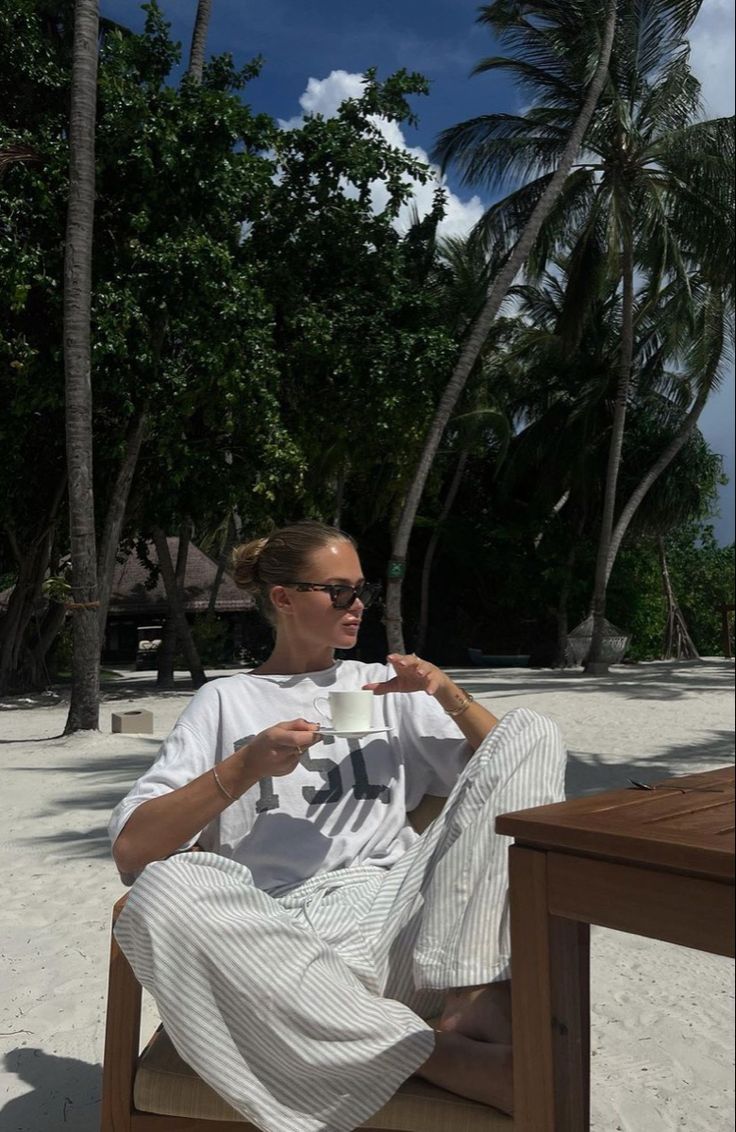 a man sitting at a table on the beach drinking from a cup with his hands