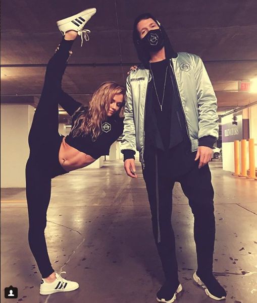 two people standing in an empty parking garage, one wearing a face mask and the other holding a pair of sneakers